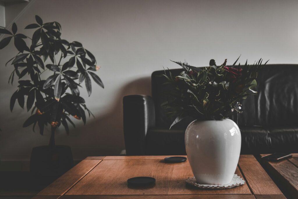A vase of flowers on a wooden coffee table beside a sofa and leafy plants.
