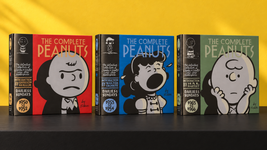 Three Complete Peanuts hardcovers—1950–1952, 1953–1954 and 1965–1966—standing side by side against a yellow wall on a black surface.