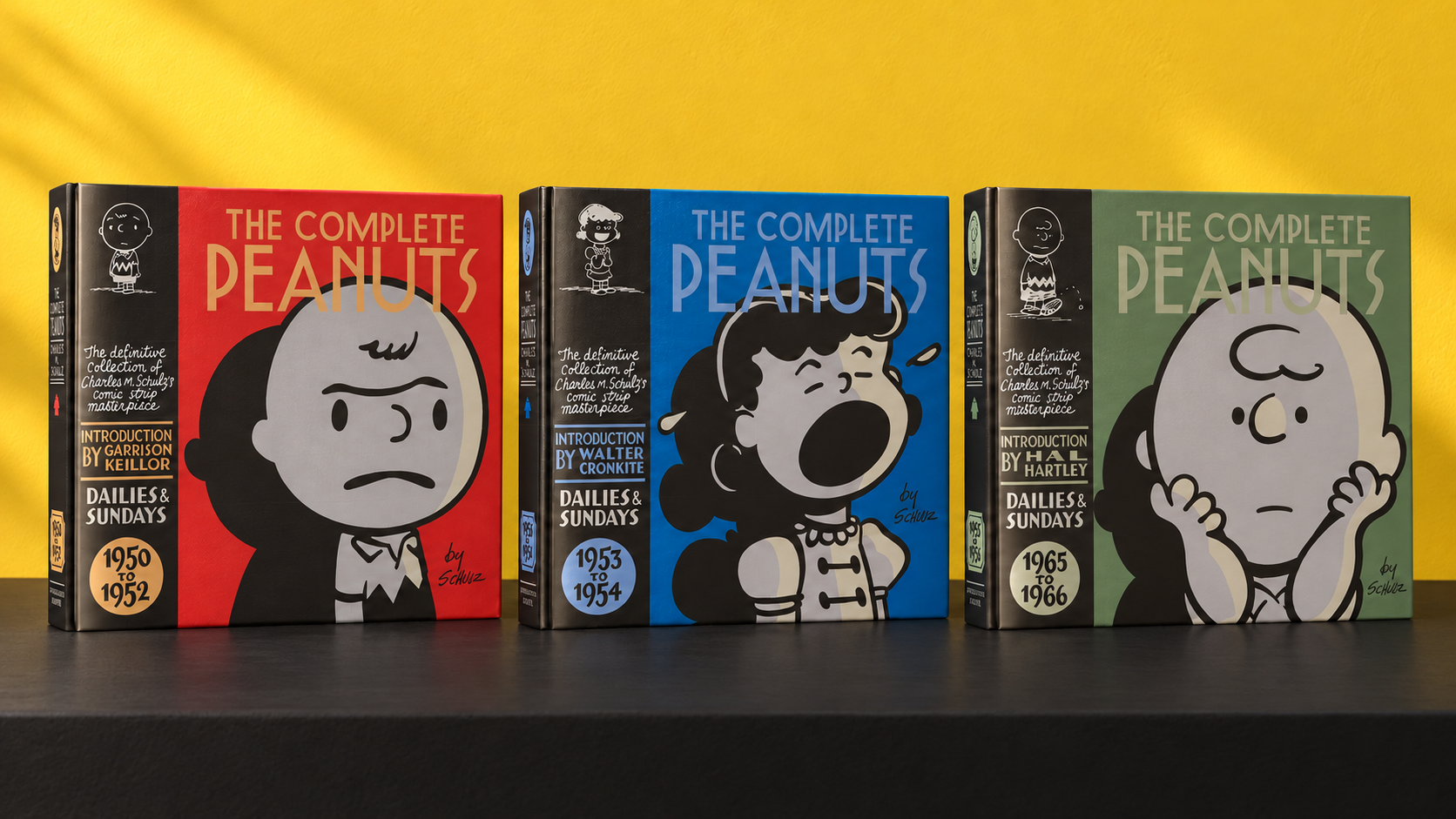 Three Complete Peanuts hardcovers—1950–1952, 1953–1954 and 1965–1966—standing side by side against a yellow wall on a black surface.